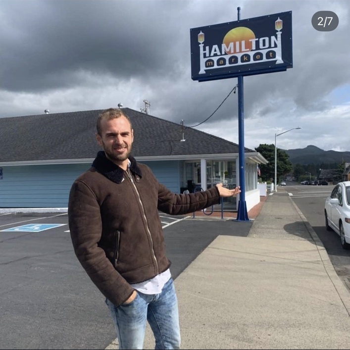 Nathanael outside the former family grocery store at 250 Avenue U in Seaside, photographed with the Hamilton Market sign.