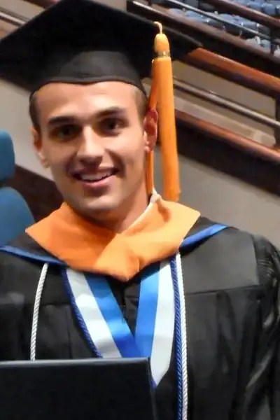 Nathanael smiling in cap, gown, and nursing hood, holding his Pensacola Christian College diploma on graduation day.